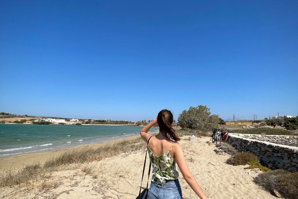 a girl facing backwards standing on a wild sand beach in paros
