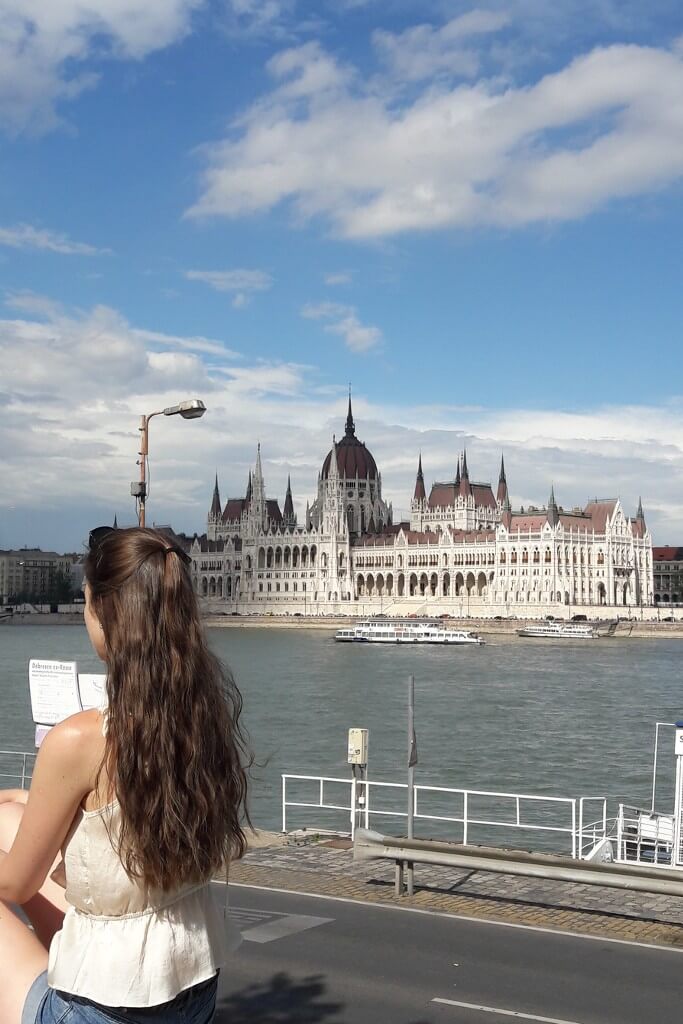 natali sitting on a low stone wall across the river from budapest parliament buidling