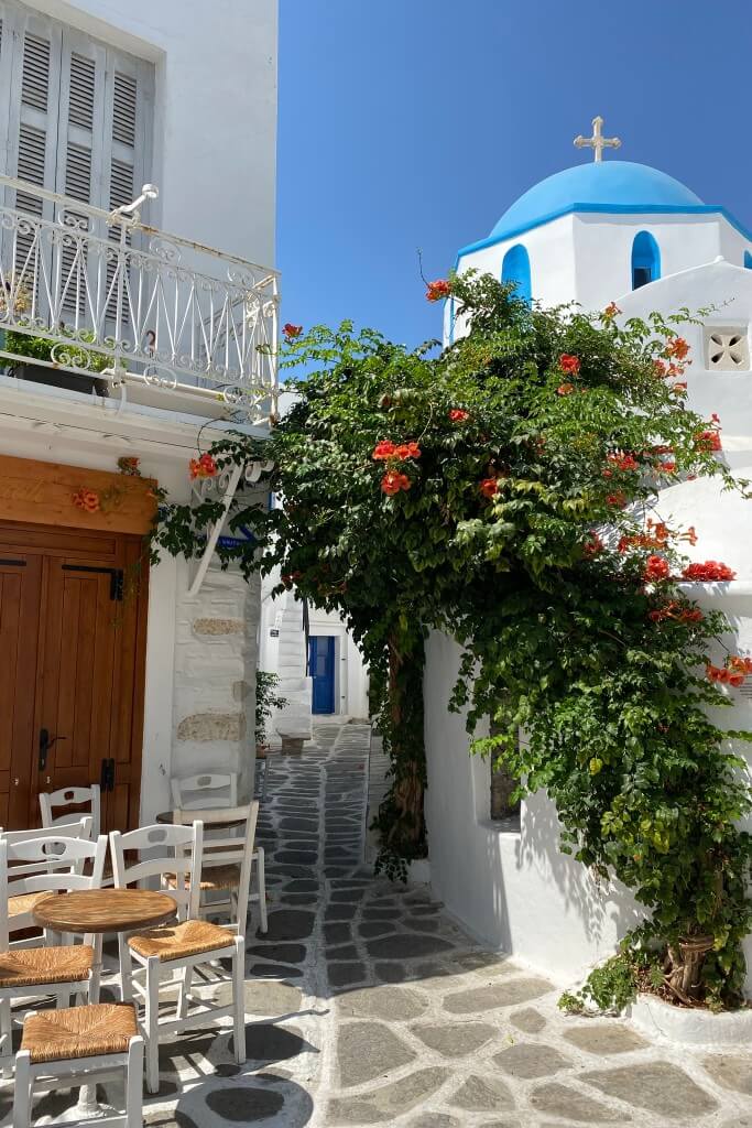 marble paved street in the city center of parikia with a white church with blue dome on one side and a white buidling with wooden chairs in front of it on the other