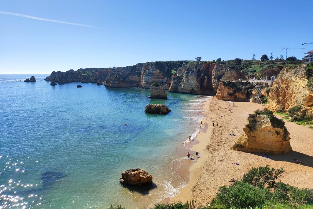 large sand beach in lagos portugal praia de dona ana, it has a single tall cliff in the middle of the beach ad it is surrounded by tall cliffs