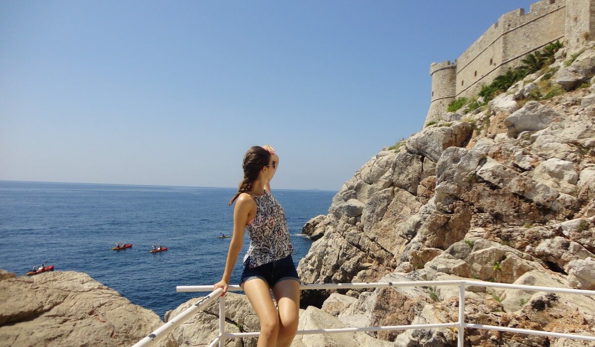 a girl sitting on a metal fence overlooking dubrovnik city walls with the sea in the background