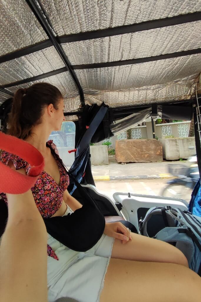 girl wearing hite whorts and flower top sitting in the back seat of a tuk tuk traveling solo in cambodia, looking through the window at the street passing by