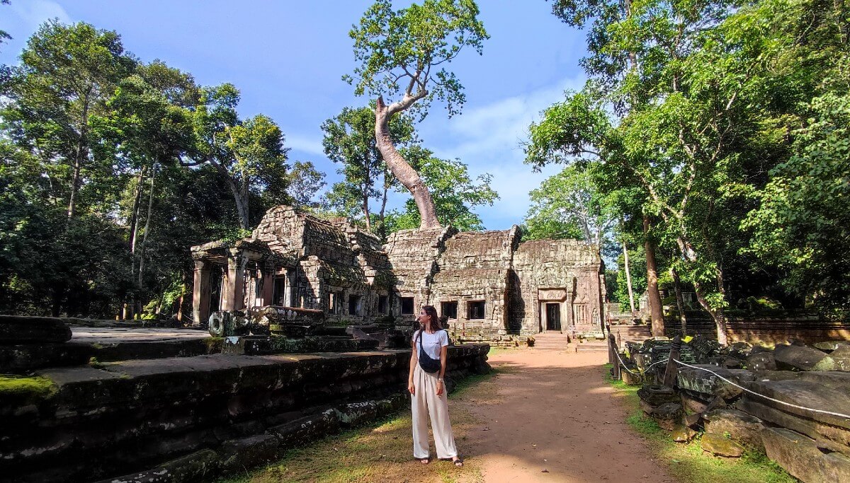 a girl wearing white pants and shirt standng in front of a temple in cambodia with a tree growing from the temple