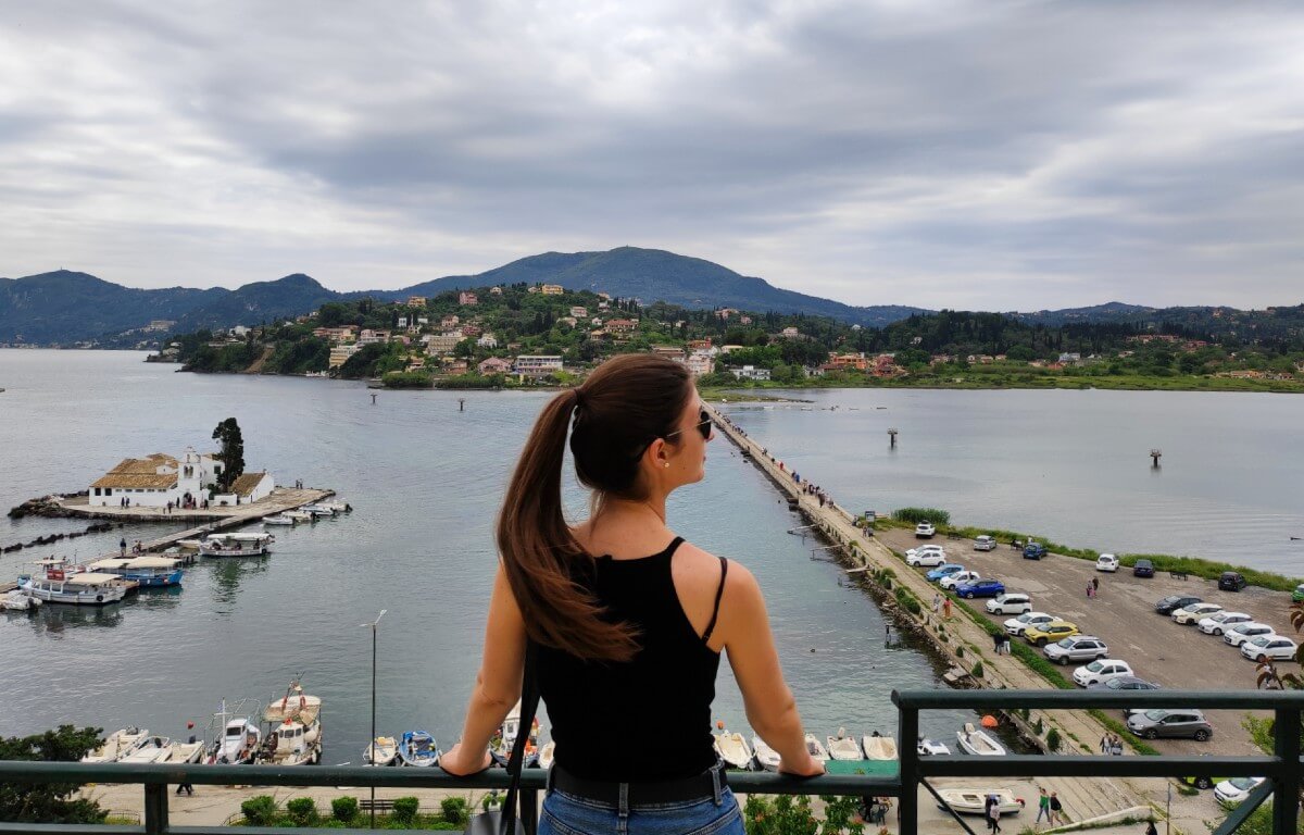 natali standing on a viewpoint in corfu in may with old town Vlacherna Monastery in front of her