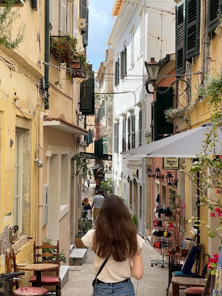 natali walking away from the camera in a narrow street in corfu town lined with souvenir shops and colorful buildings