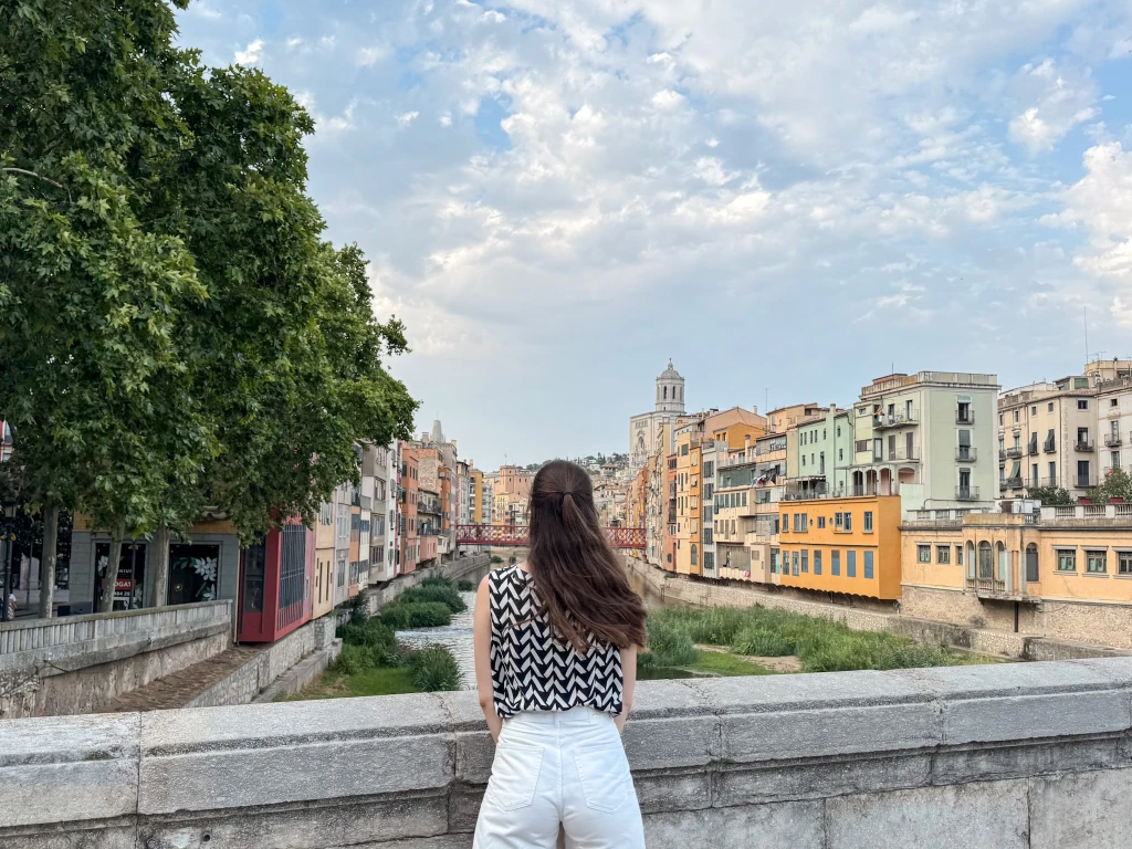natali facing away from the camera looking over the riverside of girona with colorful houses lining the river on a clear summer day